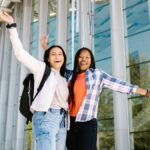 Two happy college students with raised hands posing in front of a glass building, exuding joy and friendship.