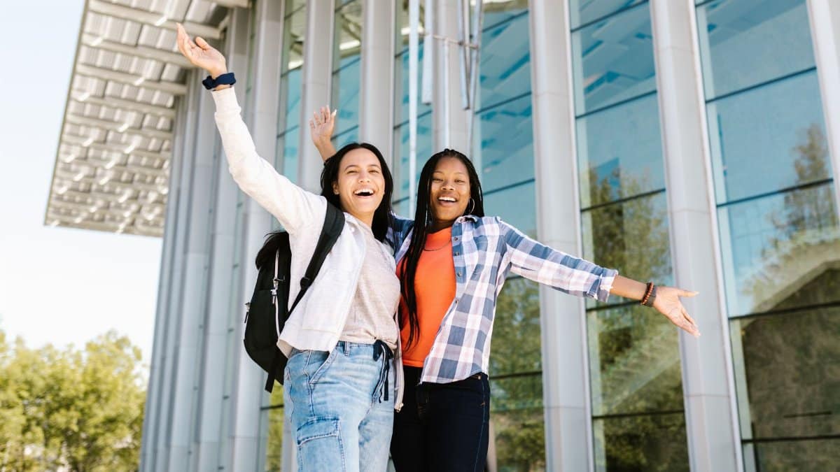 Two happy college students with raised hands posing in front of a glass building, exuding joy and friendship.