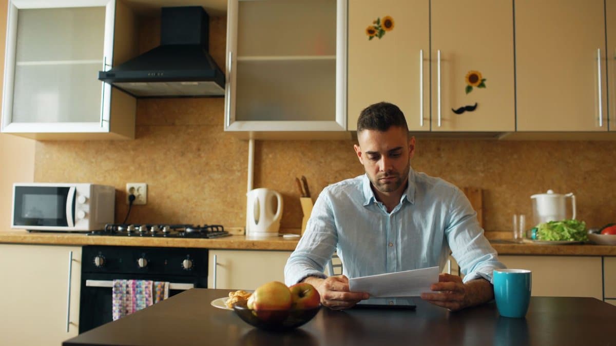 Young adult man reading at a kitchen table surrounded by modern appliances and decor.