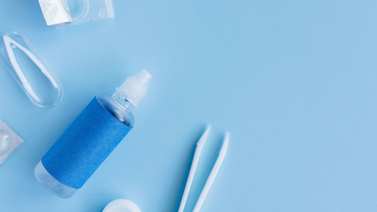 Flatlay of contact lens solution, case, and tweezers on blue background for eye care essentials.
