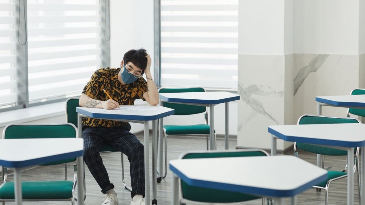 A lone student wearing a mask writes at a desk in an empty classroom following COVID-19 safety measures.