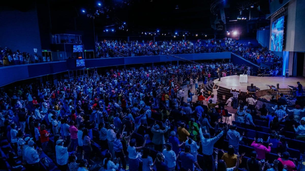A large vibrant crowd enjoying a live performance in an indoor auditorium under blue lighting.
