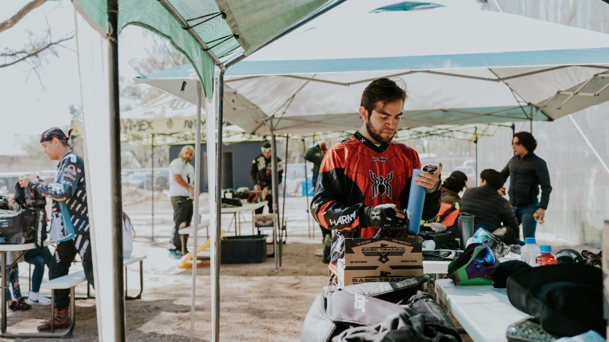 Group of adult men preparing gear for paintball under outdoor tents in daytime.