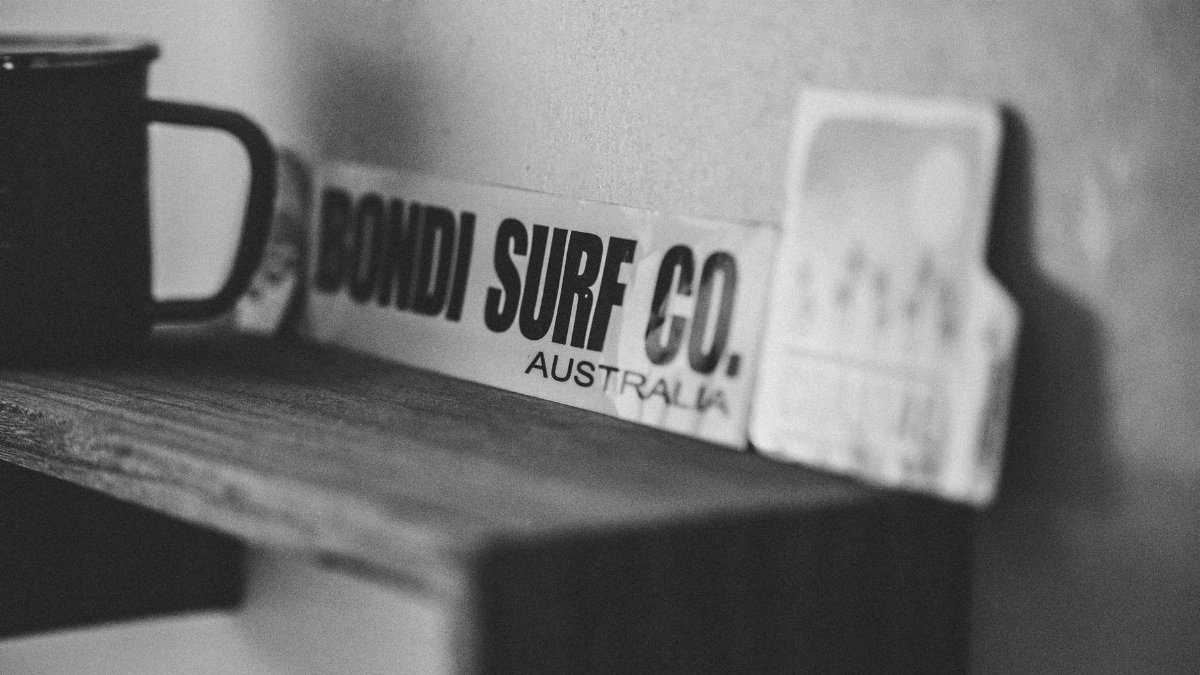 Black and white photo of a Bondi Surf Co board on a wooden shelf with a mug.