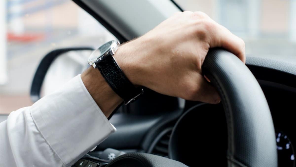 Close-up of a hand with a wristwatch gripping a car steering wheel, showcasing driving focus.