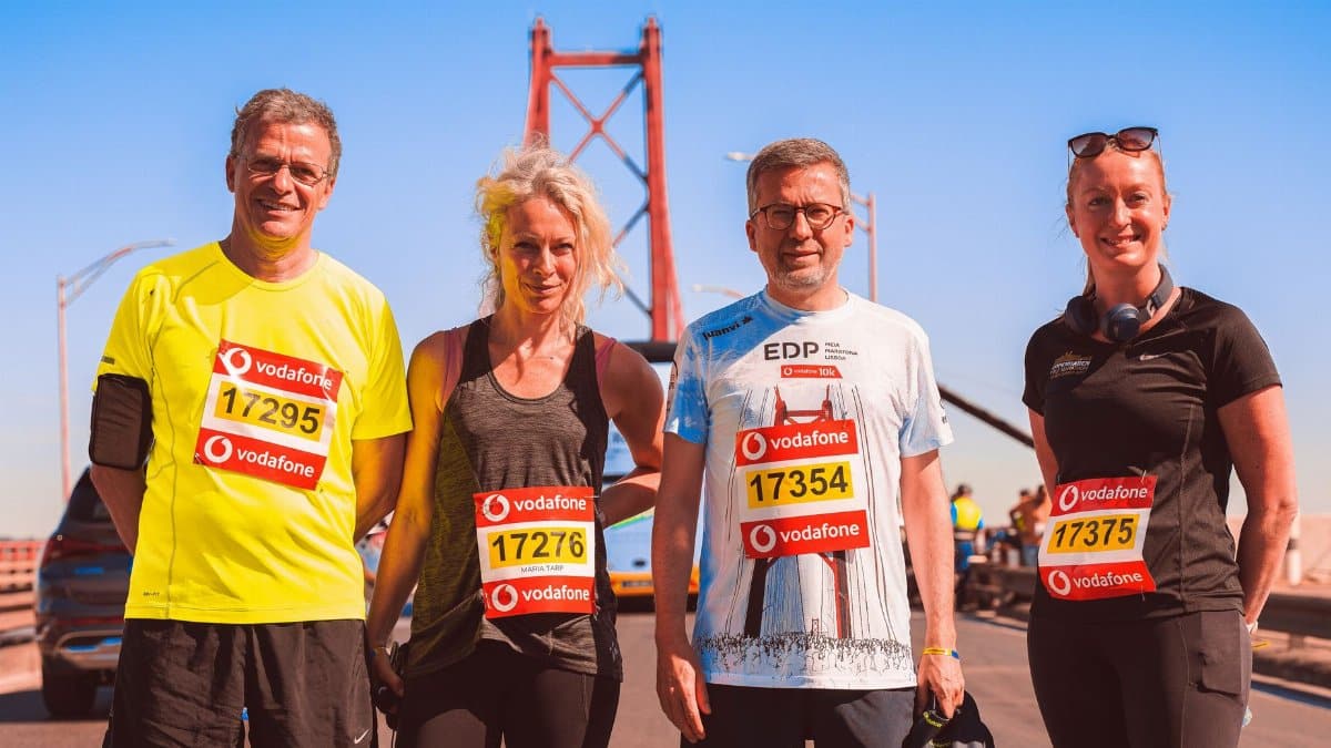 Four marathon runners smiling on a sunny day with a bridge in the background.