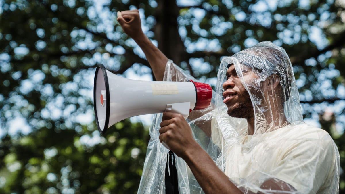 A man wearing a raincoat raises a fist and uses a megaphone to protest outdoors.