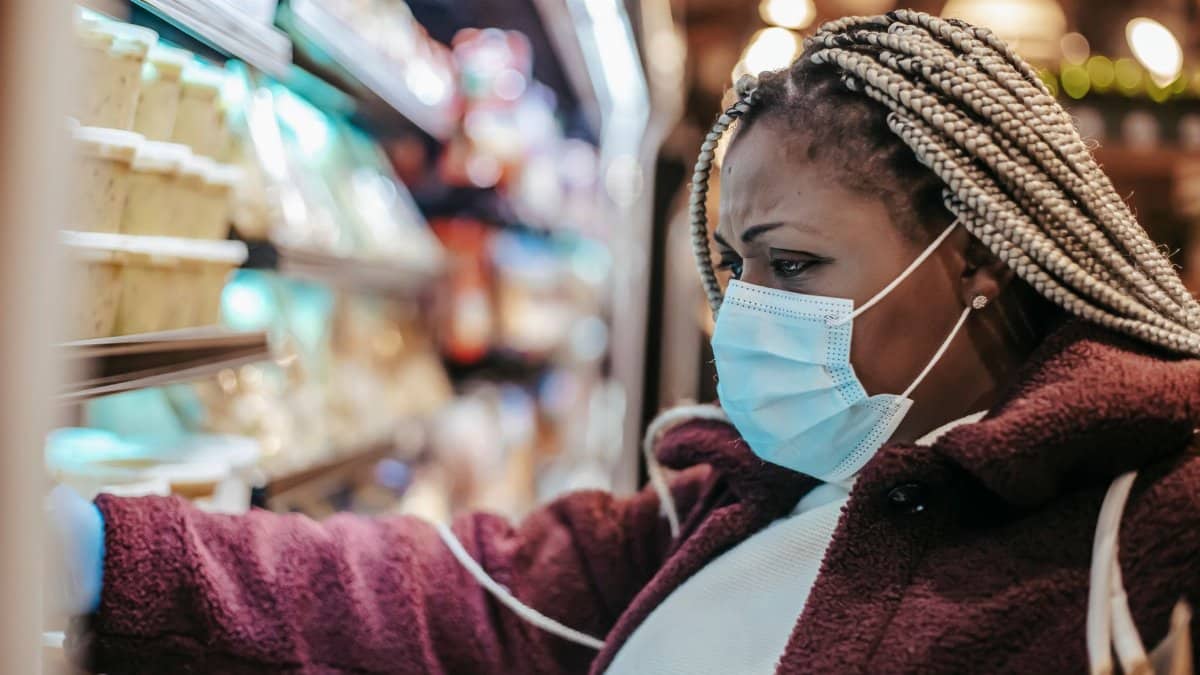 Side view of African American female buyer in medical mask picking groceries from shelf while shopping in store