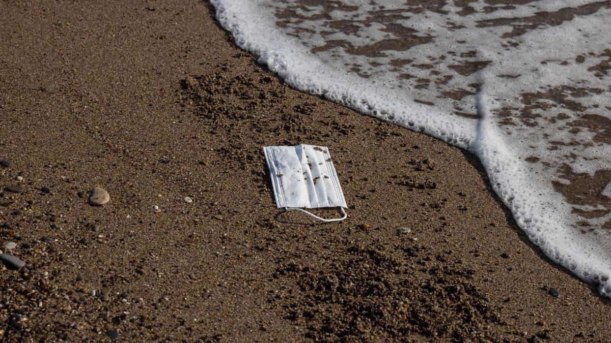 A discarded face mask lies on a beach as waves approach, highlighting environmental pollution.