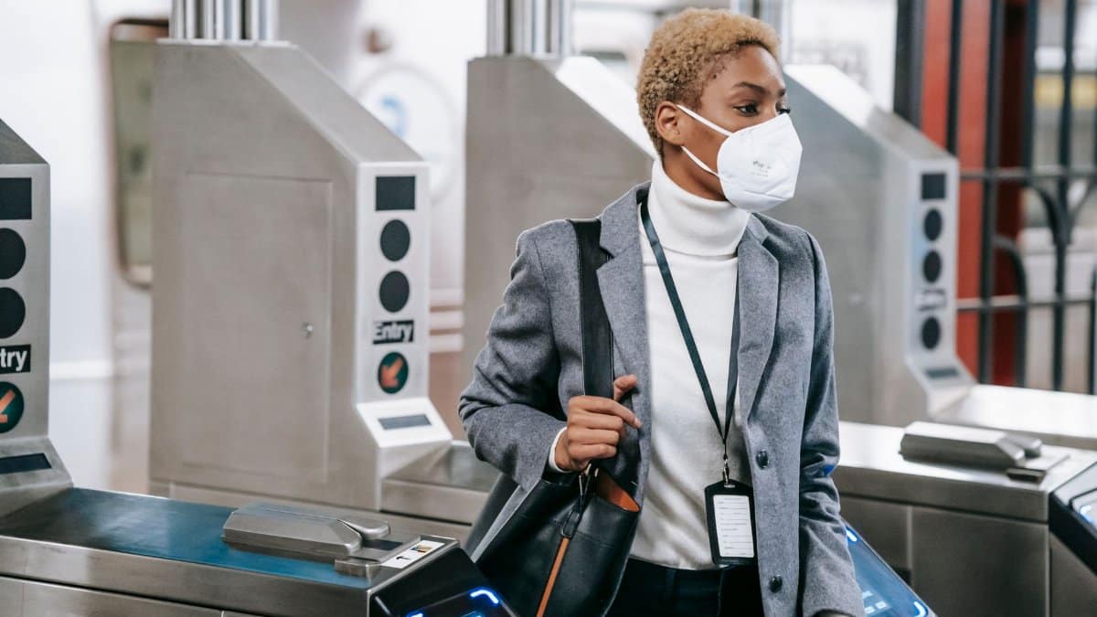Calm attractive African American female in stylish formal wear and protective mask passing through turnstile gates in underground station