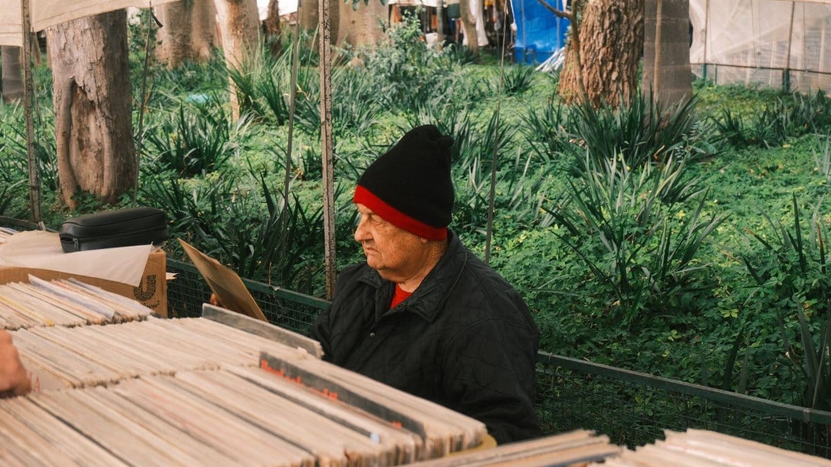 An elderly man wearing a beanie browses vinyl records at an outdoor market surrounded by trees.