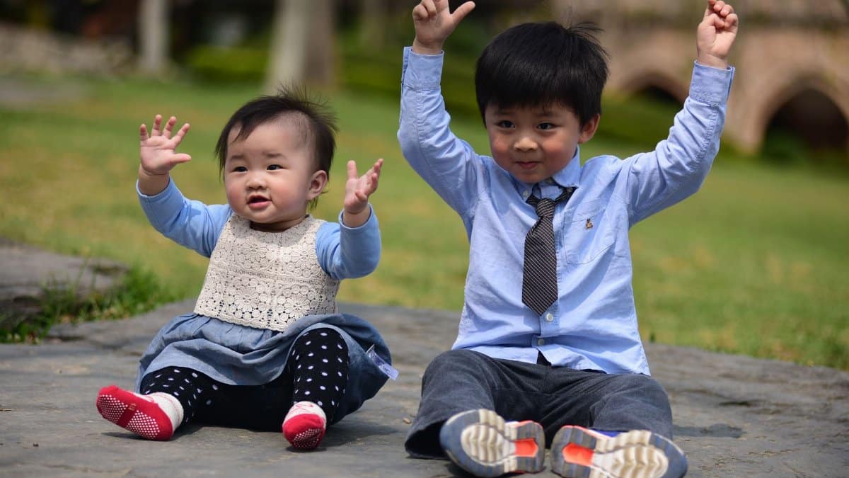 Two happy young children sitting on the ground with arms up, enjoying a sunny day outdoors.