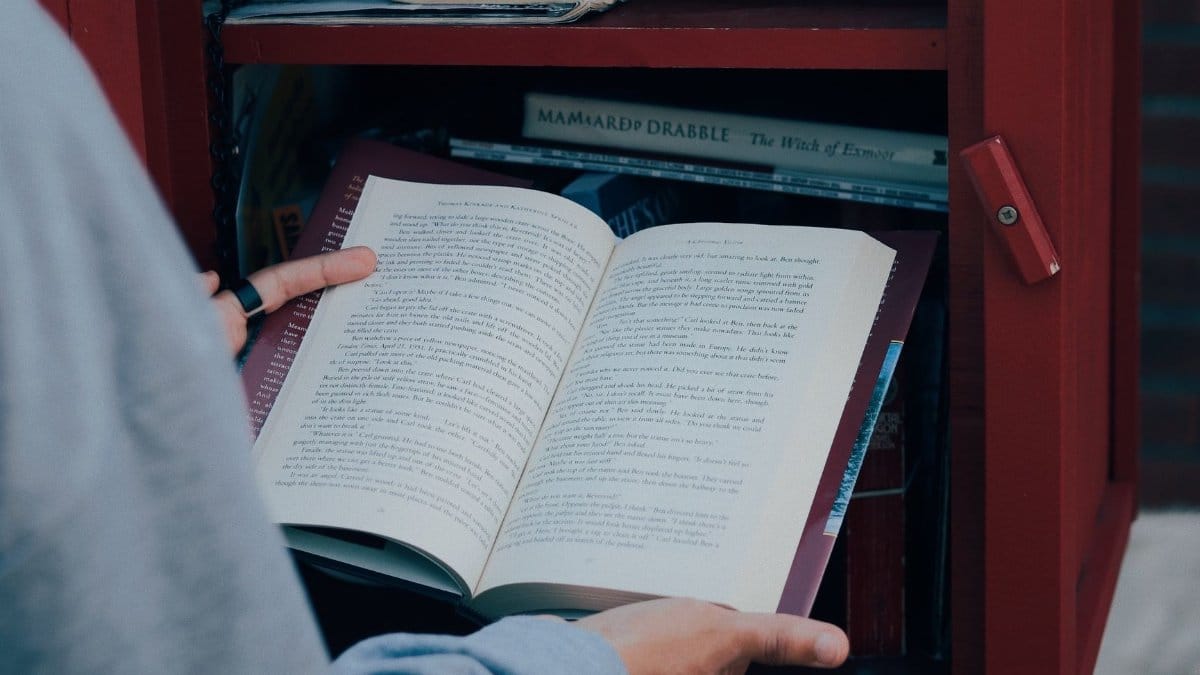 A person reads a book outside at a small free library, books visible on a wooden shelf.