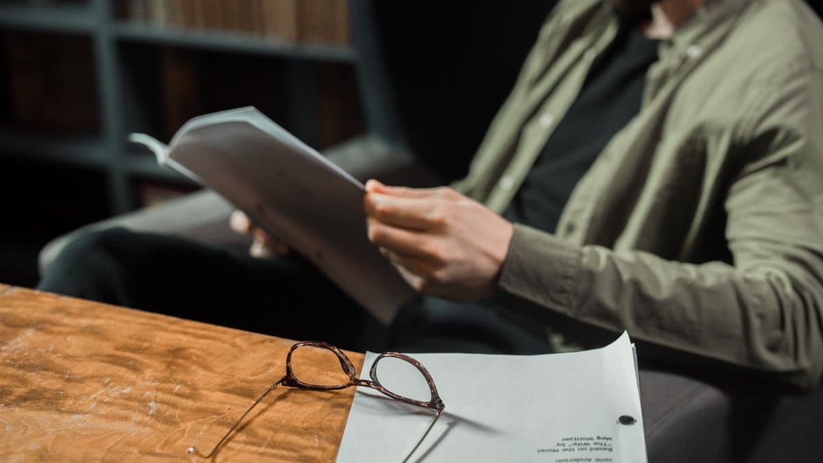A man reading in a library with glasses and papers on a wooden table.