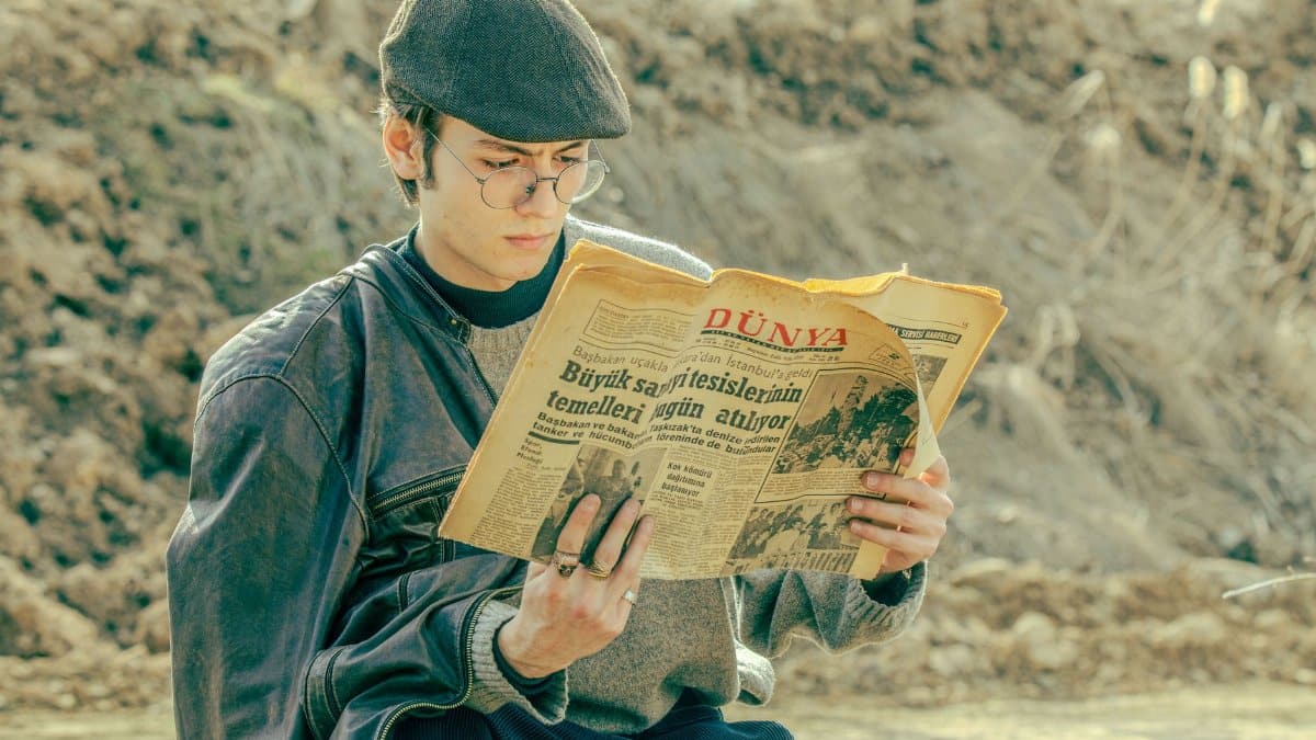 Stylish man in vintage attire reading a newspaper outdoors, capturing a serene moment.