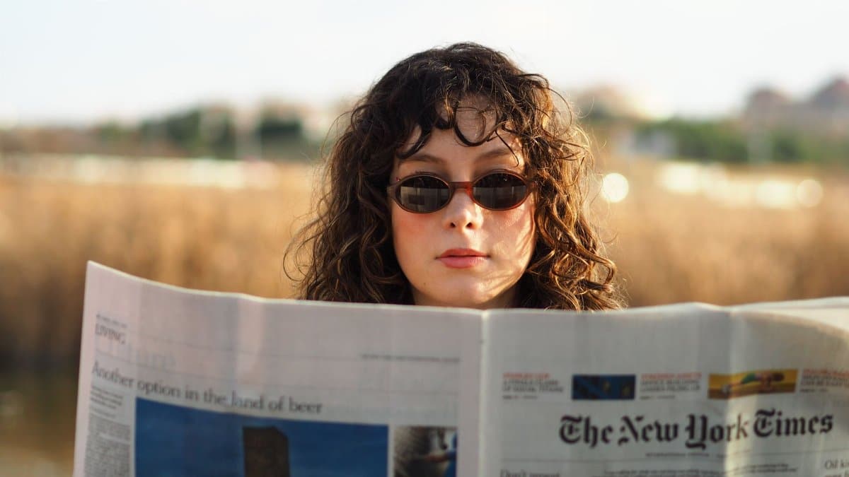 Young woman wearing sunglasses reading a newspaper in a natural outdoor setting.