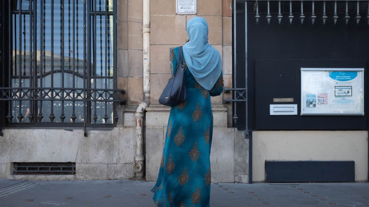 A woman in a blue dress and hijab reads a wall notice on an urban street.