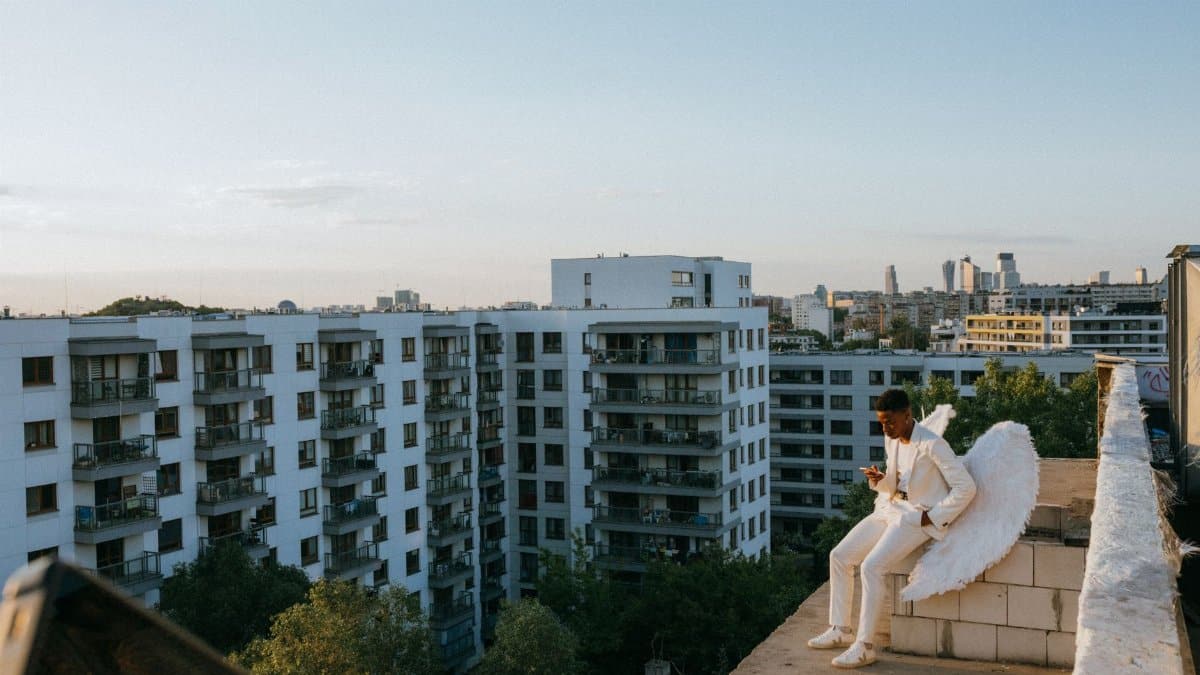 A serene scene of a man in an angel costume, sitting on a rooftop with a city backdrop.