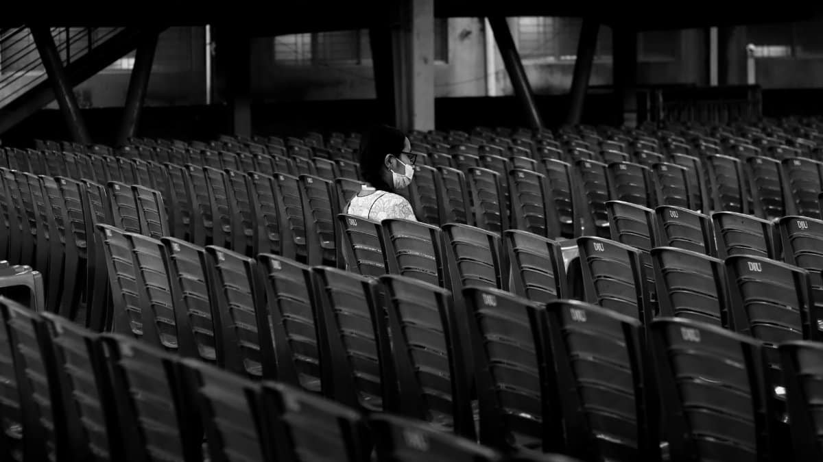 A lone person wearing a mask sits in an empty stadium, highlighting isolation during COVID-19.