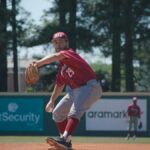 A focused baseball pitcher in red uniform prepares to throw on a sunny outdoor field.