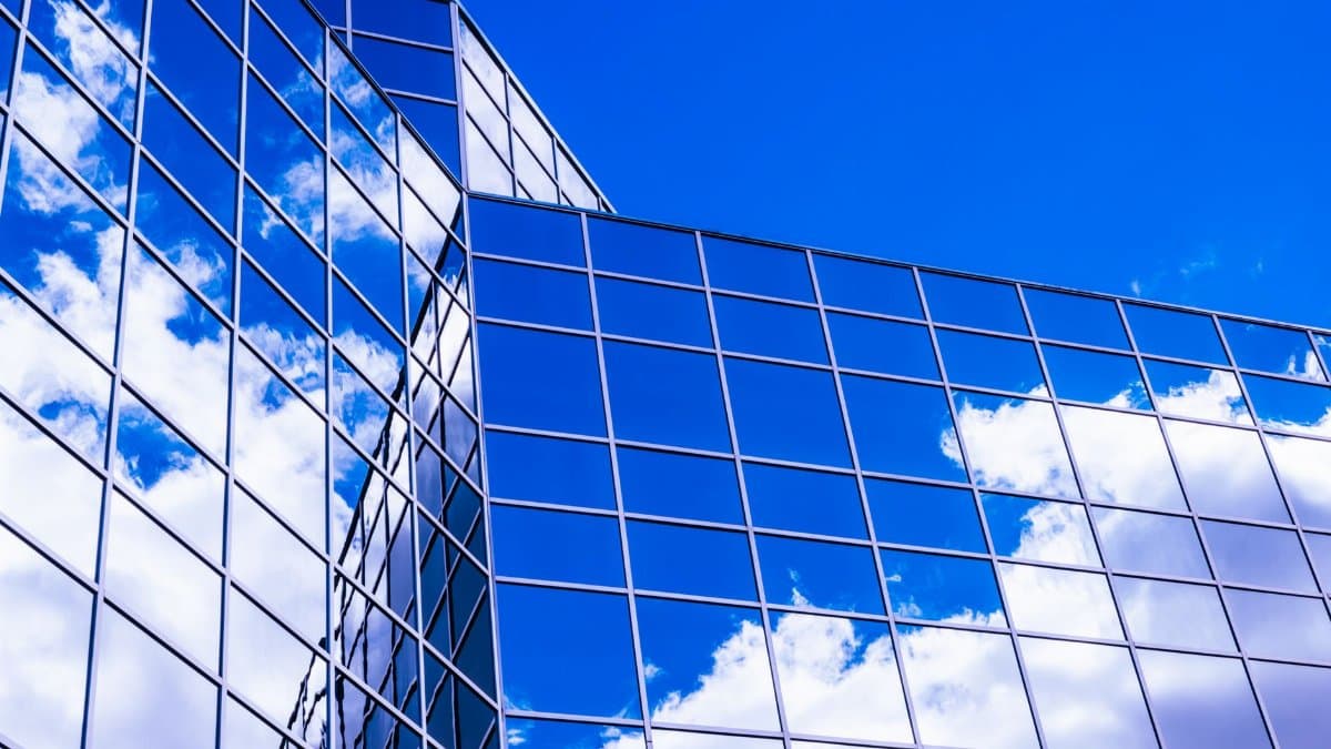 Low angle view of a modern glass building reflecting clouds against a vivid blue sky.
