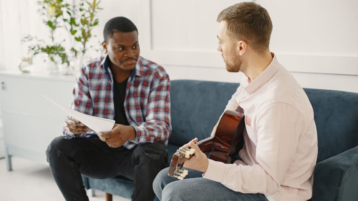 Two men sitting on a couch discussing music with guitar and papers.