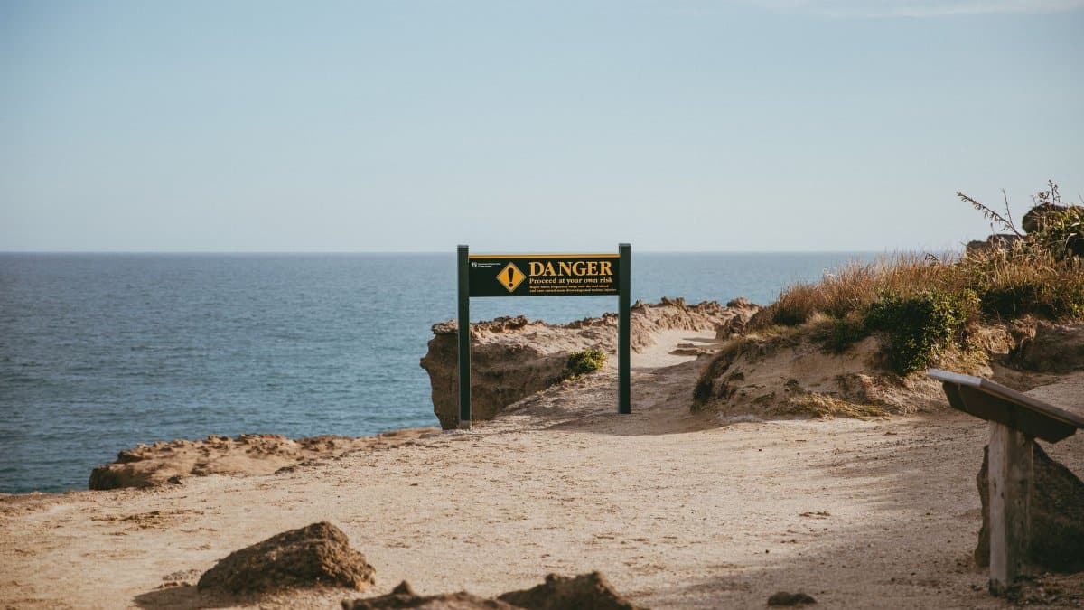 A coastal landscape in New Zealand featuring a warning sign and rugged path leading to the ocean.