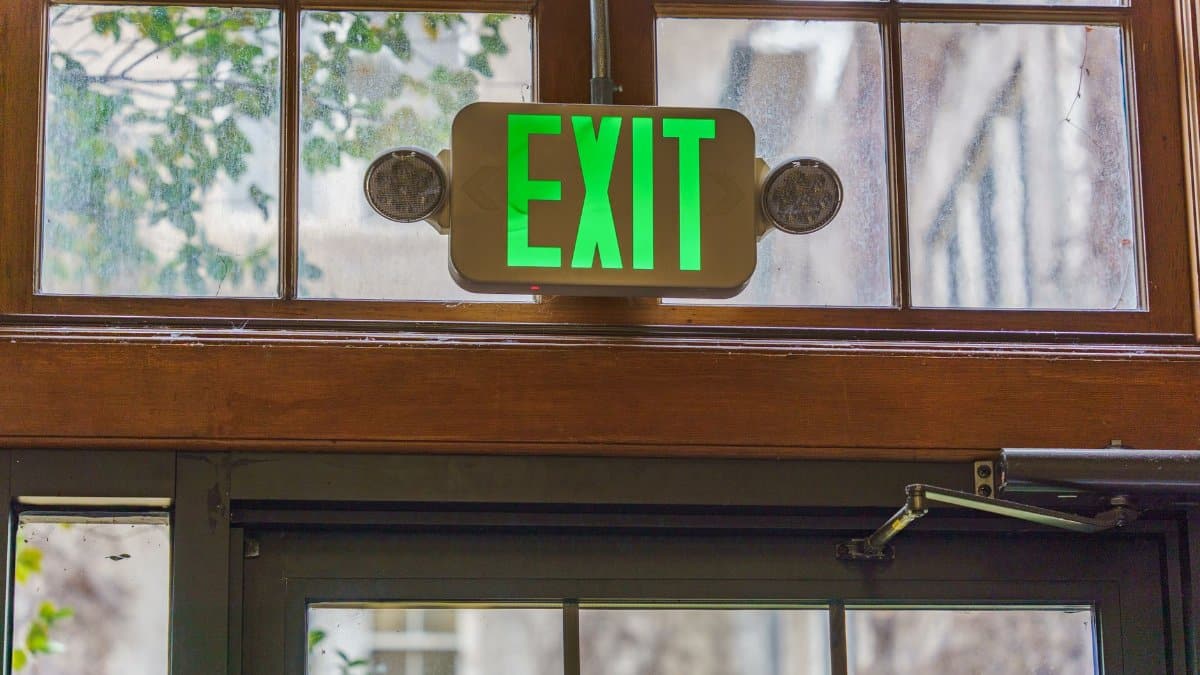 Green exit sign illuminated above a glass doorway indoors with natural light.
