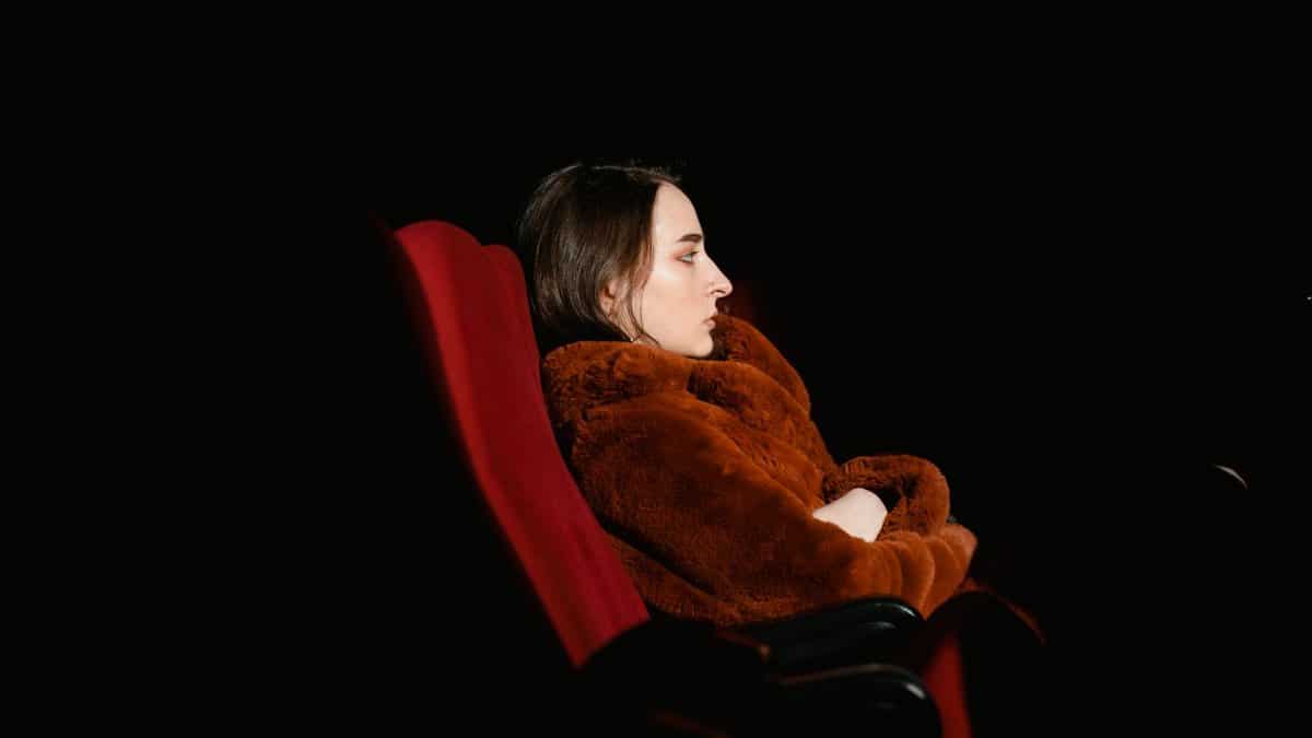 Side view of a woman in a fur coat on a red theater chair against a black background.