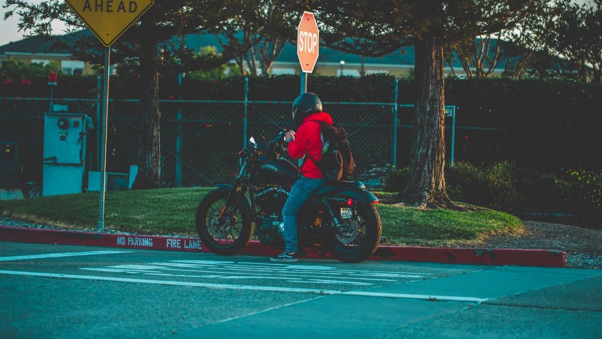 A motorcyclist in a red jacket waits at a stop sign on a urban street with traffic signage.