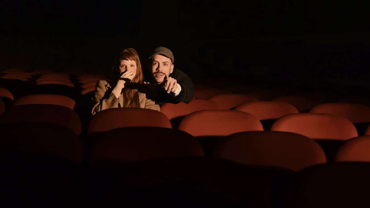 A couple sits in a dimly lit cinema theater, enjoying a movie together.