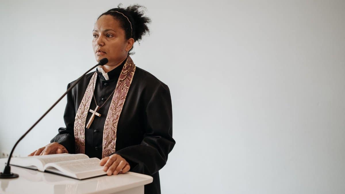 A female pastor delivers a sermon at a church podium with a Bible and microphone.