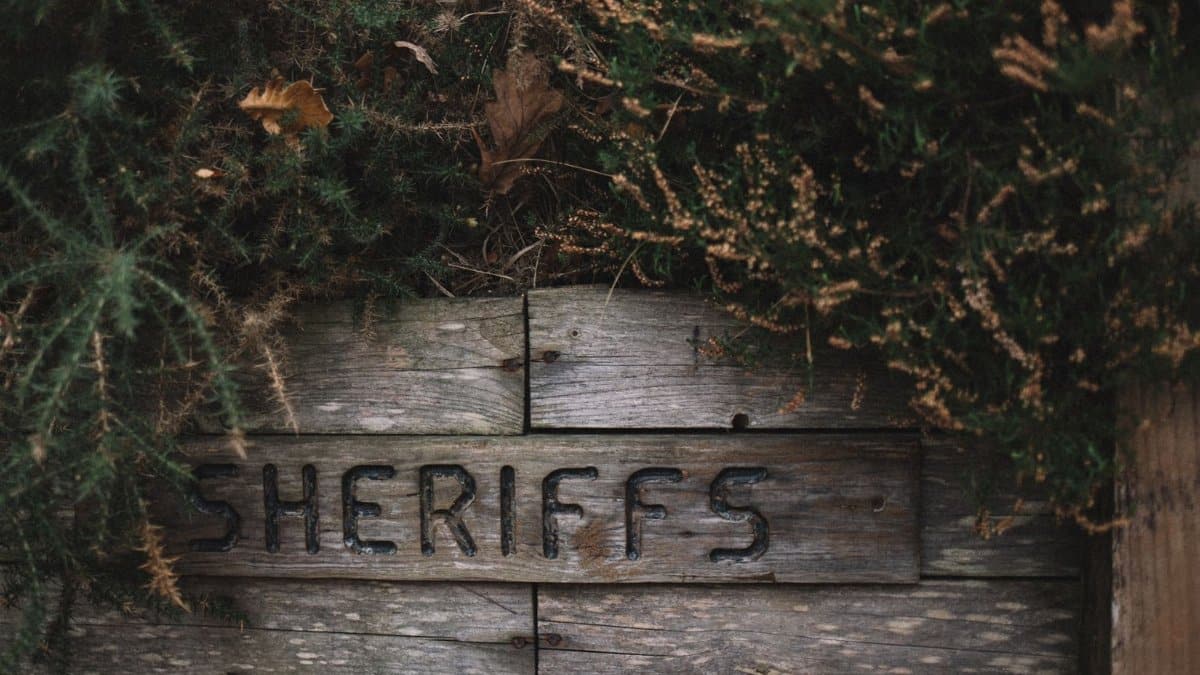 A weathered sheriff's sign on a wooden fence, surrounded by lush vegetation.