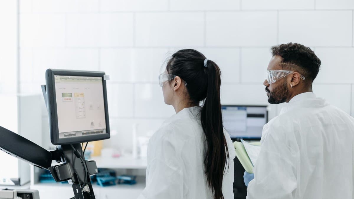 Two scientists wearing lab coats and goggles analyze data on a computer in a modern laboratory.