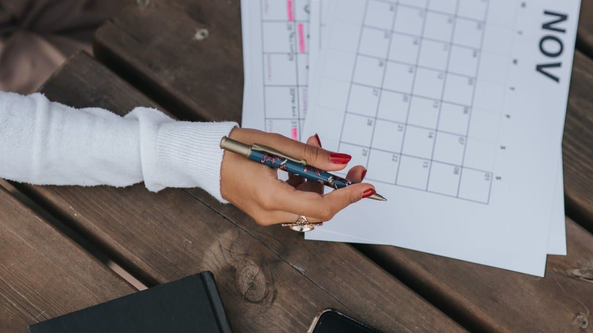 From above of crop anonymous female in white blouse sitting at wooden table with pen and calendar while making schedule