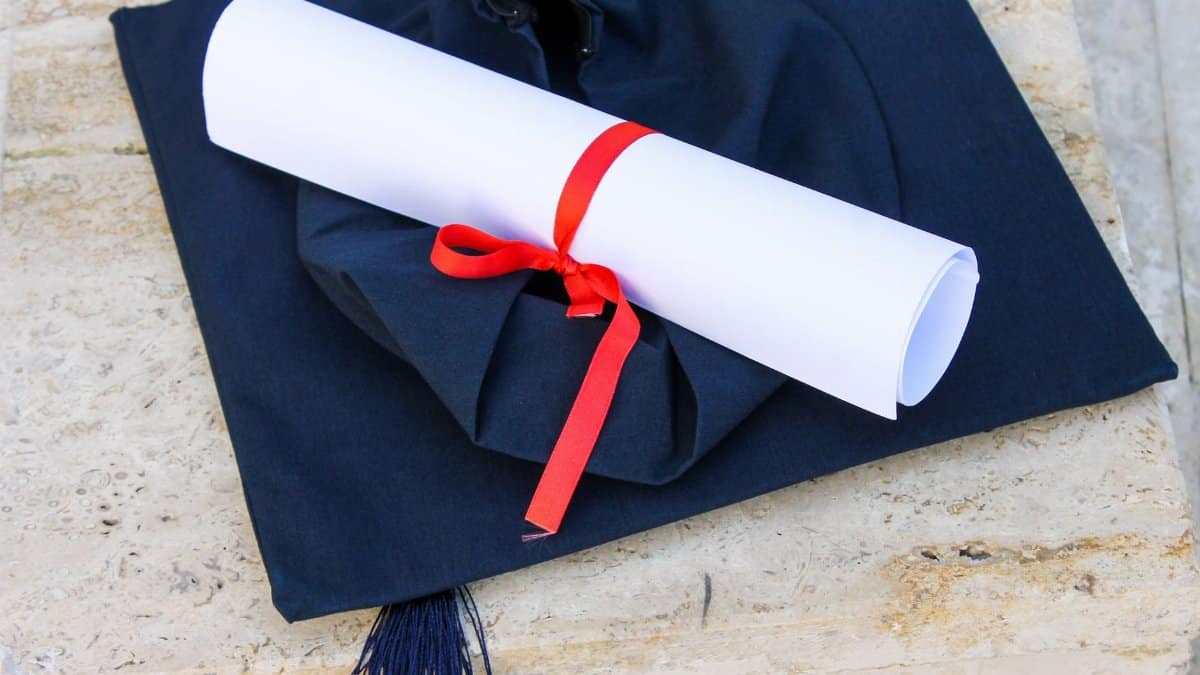 Graduation cap with diploma roll tied with red ribbon on concrete surface.