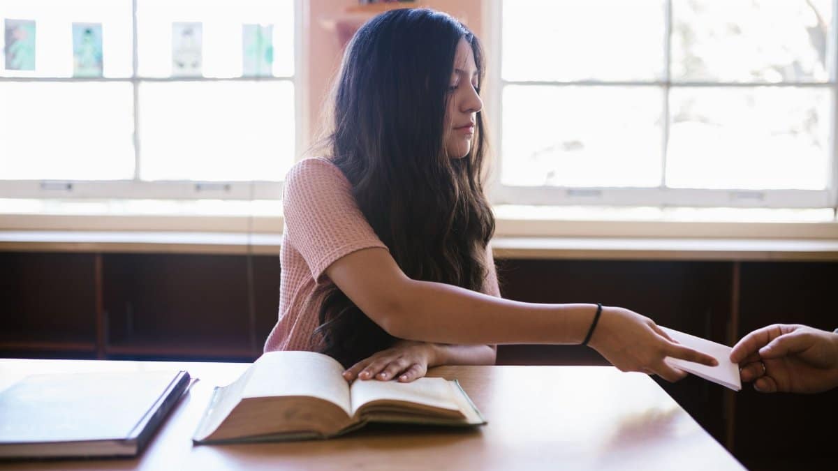 A young woman passes paper across a desk in a classroom with books and natural light.