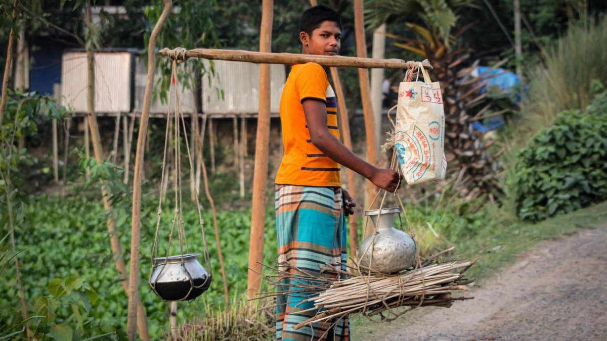 Young man carrying traditional load with pots and sack in a rural setting.