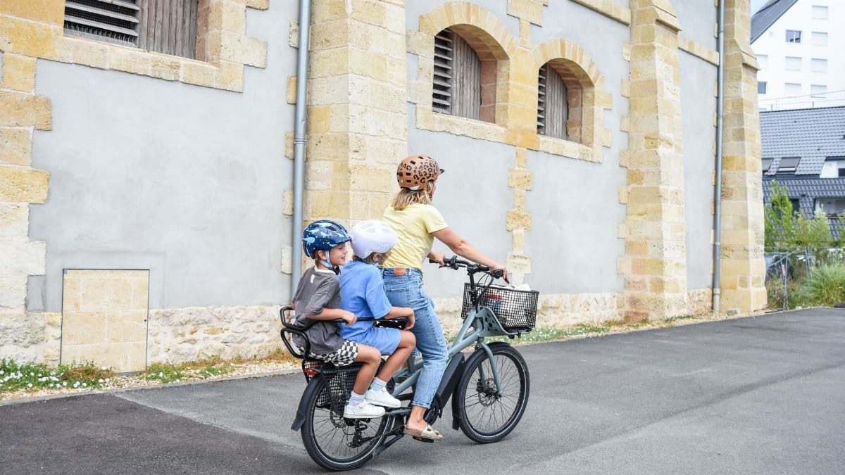 A woman and two children ride a cargo bike past a historic building on a sunny day.