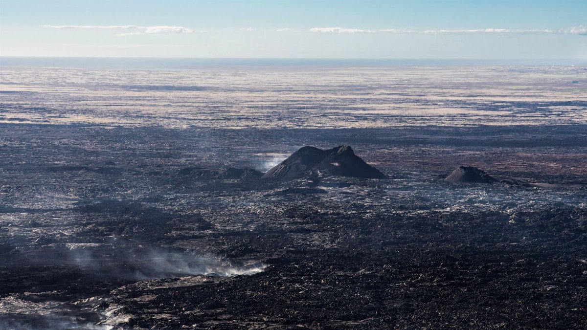 A wide aerial view of a rugged volcanic landscape with visible lava flows and smoke rising from the surface.