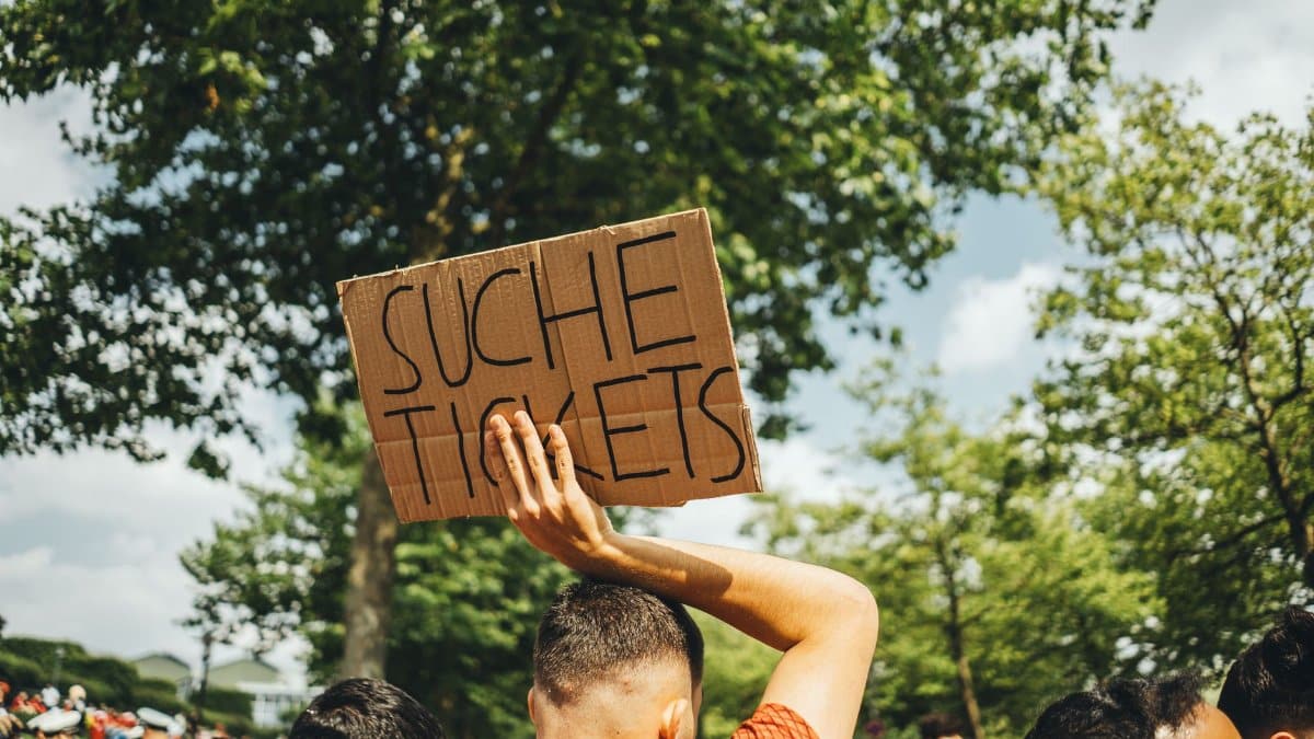 A man in a crowd holds a sign reading 'Suche Tickets' at an outdoor event, seeking extra admissions.