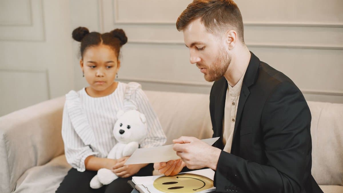 A child holding a plush toy listens to a man in a therapy session.