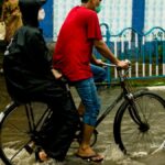 A man riding a bicycle with a passenger in a raincoat through flooded streets during heavy rain.