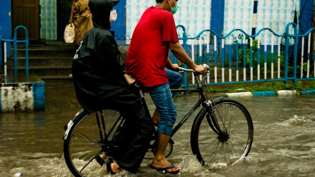 A man riding a bicycle with a passenger in a raincoat through flooded streets during heavy rain.