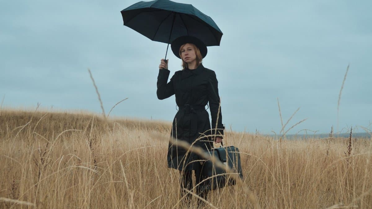 Serious young female in black coat and hat with umbrella and briefcase standing among dry grass in field and looking away in overcast weather