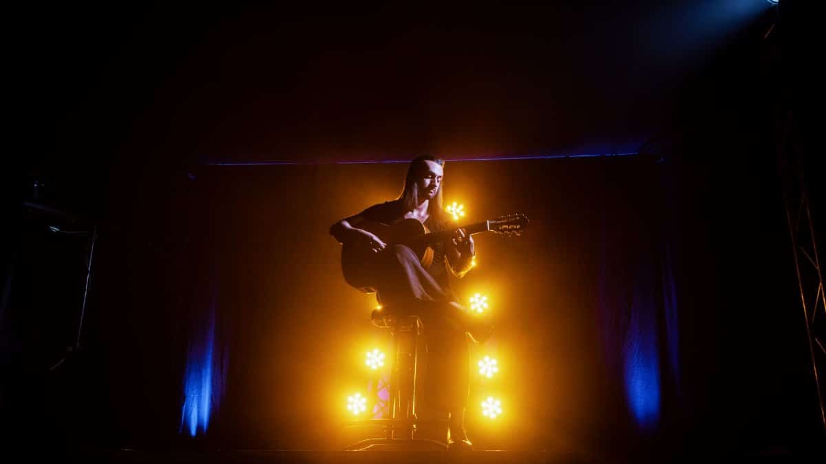 A musician playing an acoustic guitar on stage, illuminated by dramatic yellow and blue lights.