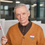 Elderly man with American flag and vote sticker in polling station. Emphasizing civic duty and participation.