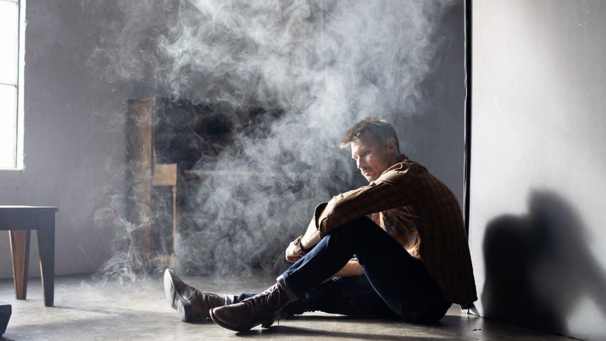 A man in boots and denim sits pensively in a smoke-filled room with soft natural light.