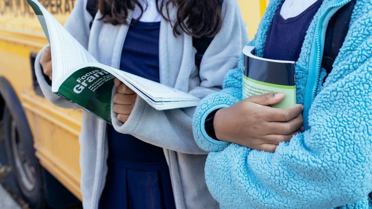 Crop faceless ethnic children holding textbooks while standing near yellow school bus together