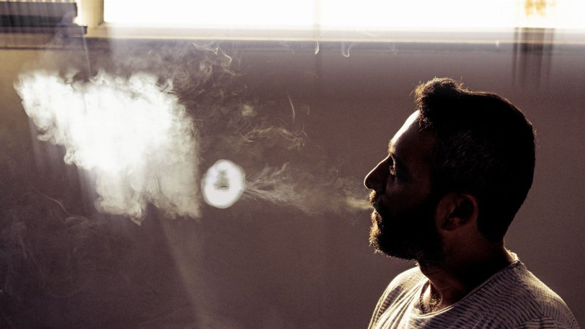 Side profile of a man blowing smoke indoors with dramatic lighting.